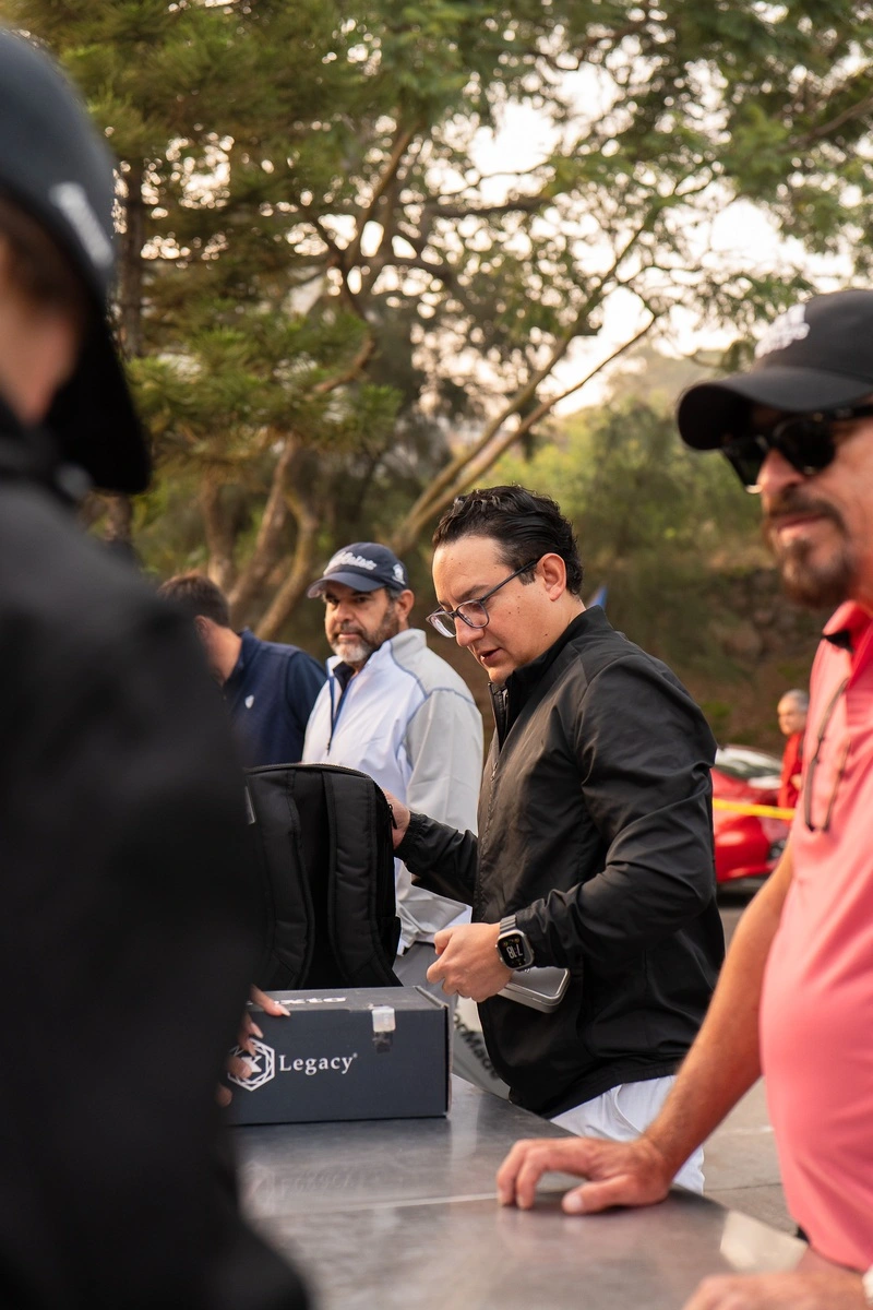 Jugador revisando su kit de bienvenida en el área de recepción del evento de golf, acompañado de otros participantes bajo un entorno arbolado