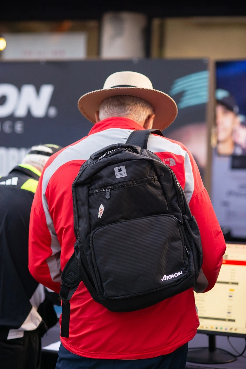 Hombre con sombrero y mochila negra de Akron visto desde atrás mientras realiza su registro en el evento, frente a módulos y pantallas informativas.