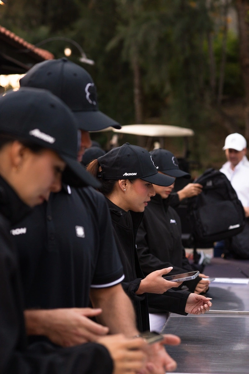 Miembros del staff alineados en el módulo de registro del evento de golf, usando gorras y chamarras negras mientras atienden a los participantes