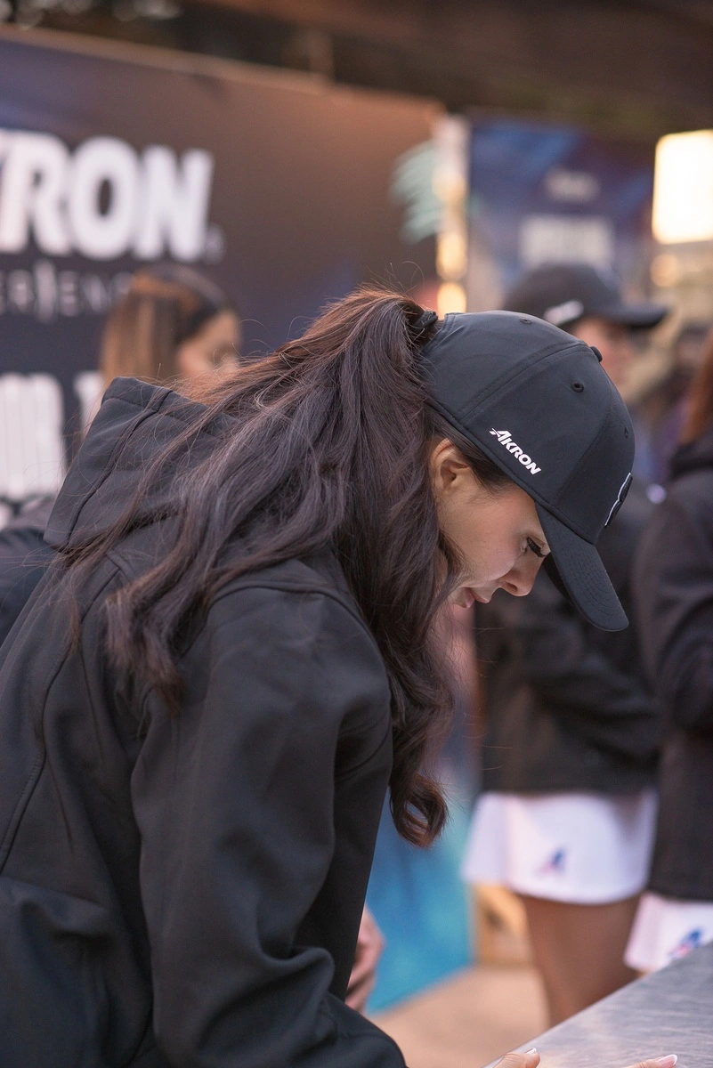 Mujer del staff inclinada mientras atiende el registro del evento, usando gorra y chamarra negras con fondo difuminado del área de bienvenida