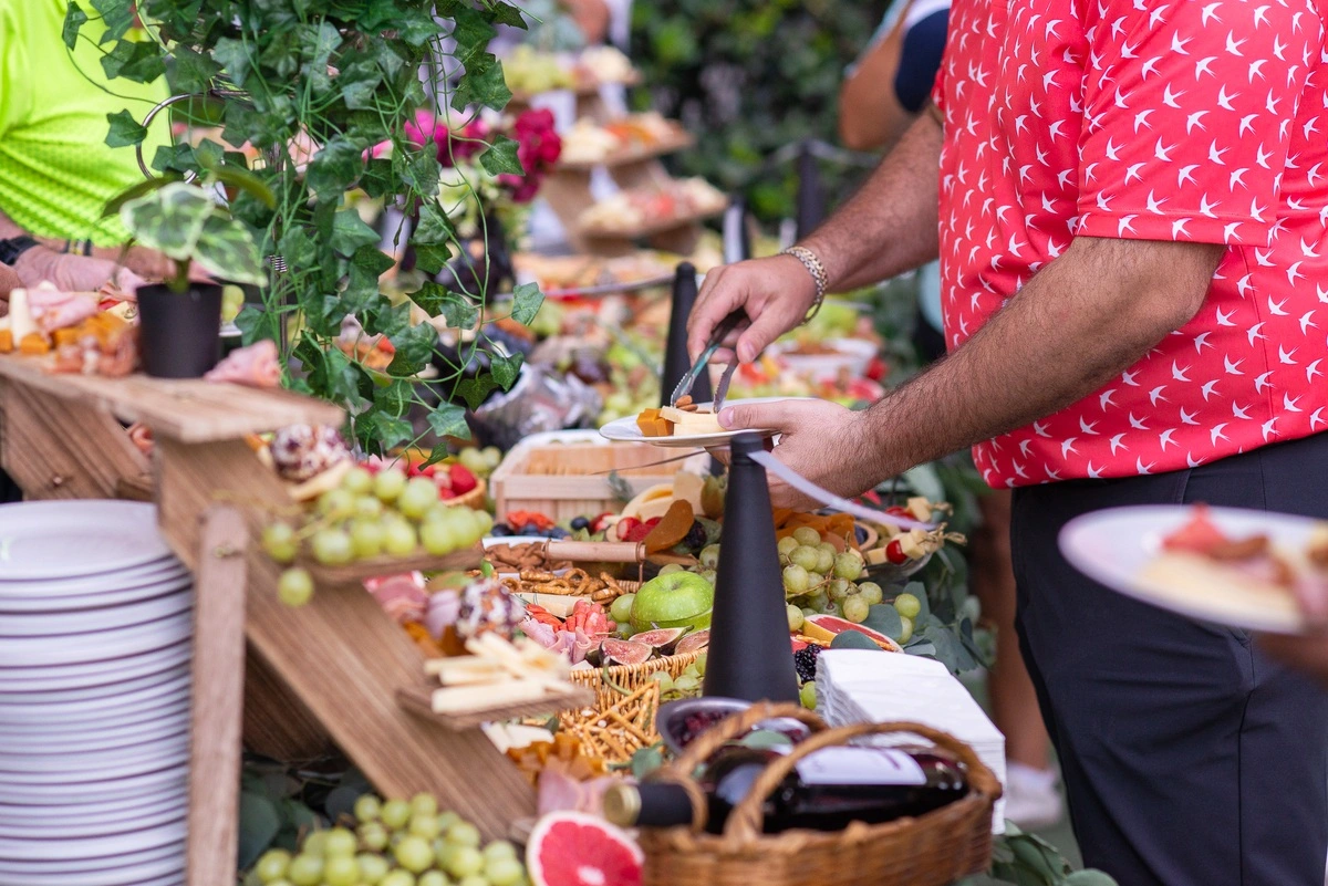 Manos de un participante sirviéndose alimentos en una mesa de buffet repleta de frutas, quesos, charcutería y bocadillos durante el evento