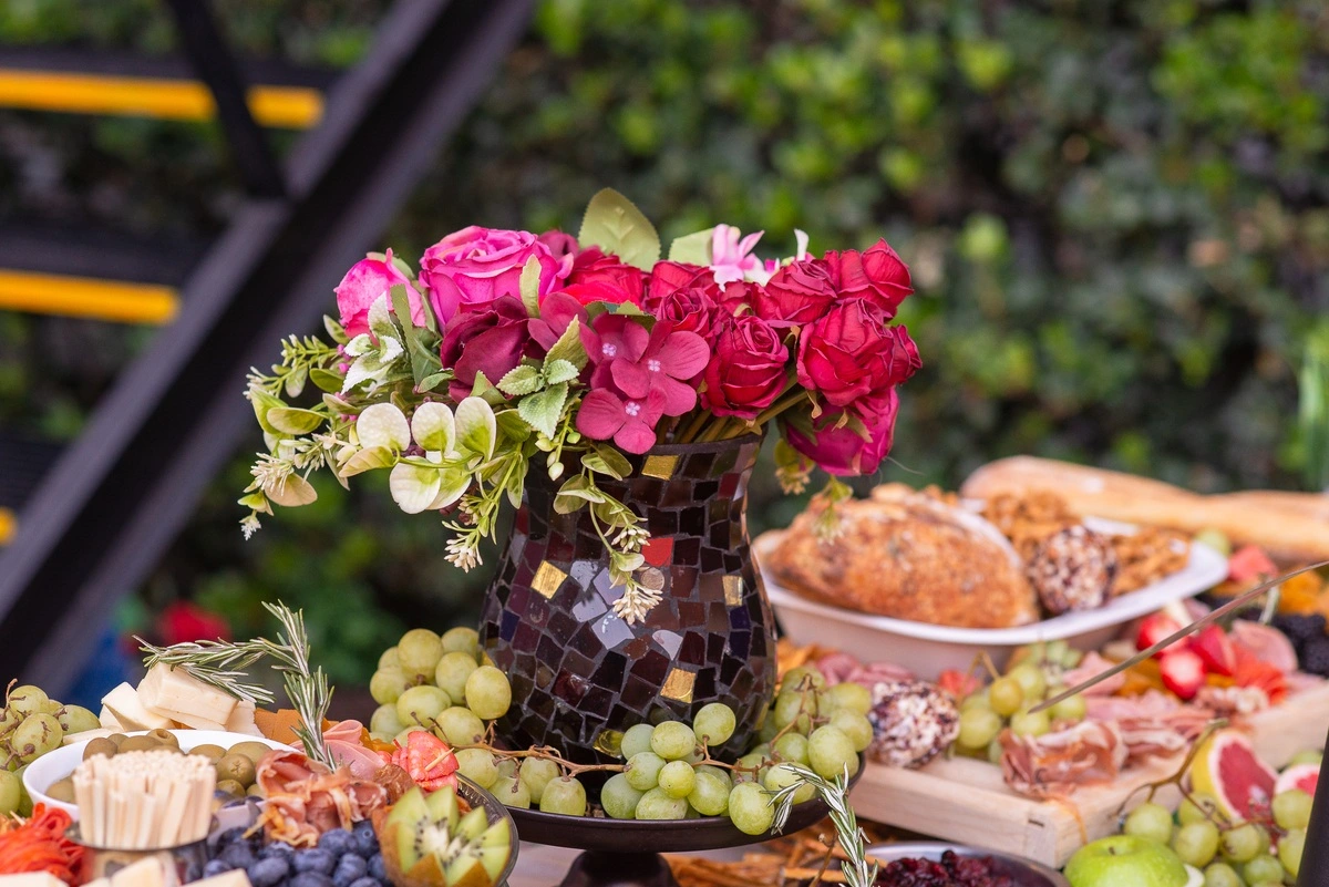 Arreglo floral en jarrón oscuro sobre mesa con frutas, quesos y charcutería en un buffet