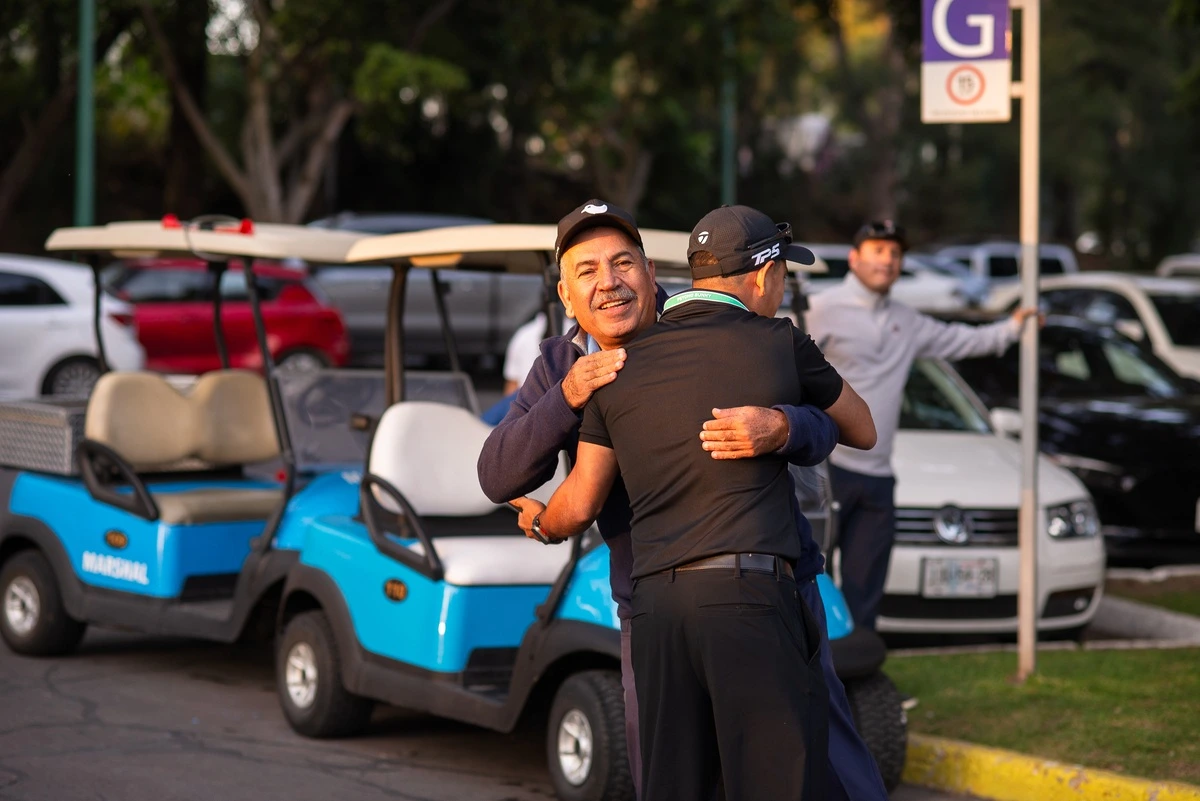 Dos participantes del torneo se abrazan sonrientes junto a carritos de golf en el estacionamiento