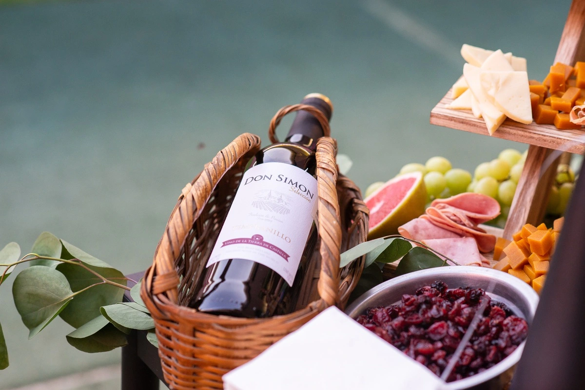 Botella de vino en canasta junto a frutas, charcutería y quesos en mesa de buffet.